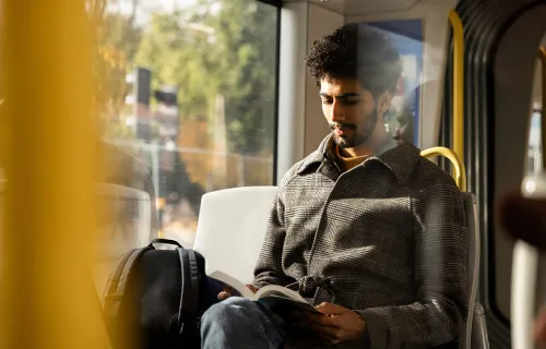 En man sitter vid fönstret i en buss eller spårvagn och läser en bok. Solljus faller in genom…