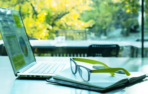 An open laptop with graphs on screen, a note pad and pair of green glasses on a table facing a sunny garden.