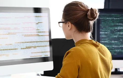 female software engineer sat at a desk looking at software on 2 laptop screens 