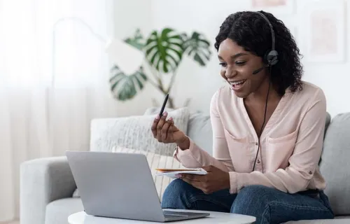 Smiling young woman sitting on sofa and talking on headset while looking at laptop