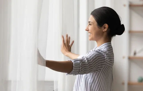 woman in white shirt opening curtains smiling