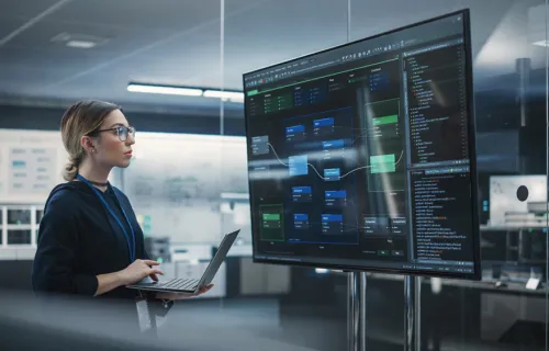 Woman holding a laptop and looking at a large monitor