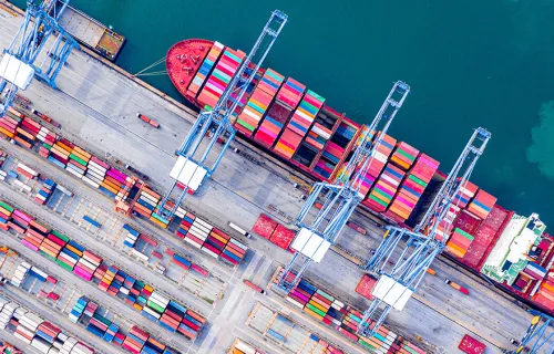 arial view of a shipping yard with a barge docked and lines of shipping containers 