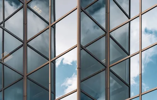Närbild av en modern byggnads glasfasad med geometriska metallramar. Blå himmel och moln…