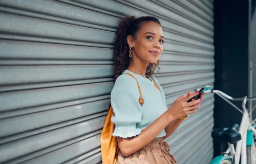 Person leaning against a metal shutter while holding a smartphone, with a bicycle and backpack…