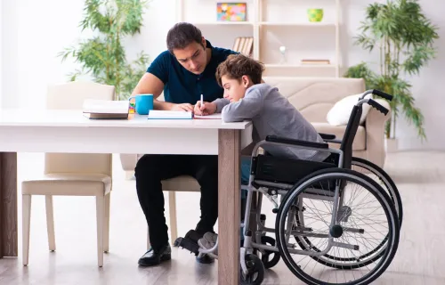 Adult helping a child in a wheelchair with schoolwork at a table in a bright, comfortable home…