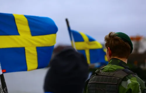 Svenska flaggor vajar i vinden medan en person i uniform står med ryggen mot kameran. Bilden…