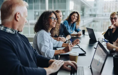 En grupp kollegor sitter runt ett långt konferensbord i ett modernt kontor med stora fönster…