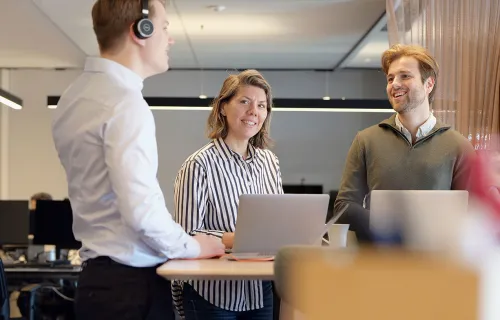 Tre kollegor har ett stående möte vid ett högt bord i ett modernt kontorslandskap. Två personer…