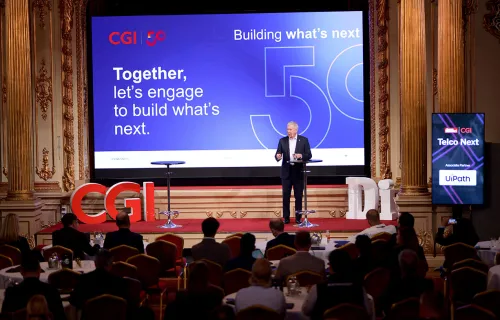 En talare står på scen under ett CGI-evenemang inför en sittande publik i en elegant…