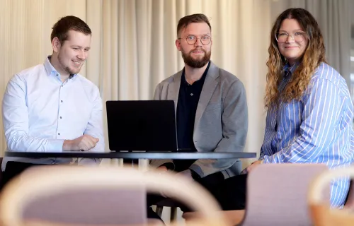 Tre kollegor sitter tillsammans vid ett bord med en laptop och samarbetar. De ler och verkar ha…