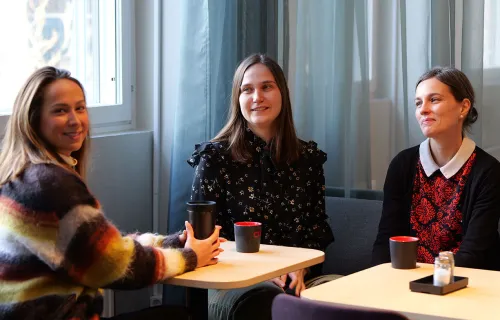 Tre kvinnor sitter vid små bord i ett ljust rum och samtalar över kaffe. De ler och ser…