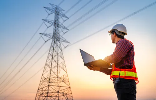 Arbeider med hjelm og refleksvest bruker laptop ved høyspentmast.