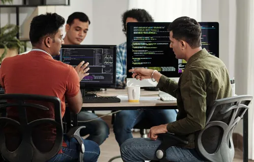 Team members having discussion at desks in front of computers with coding on screen