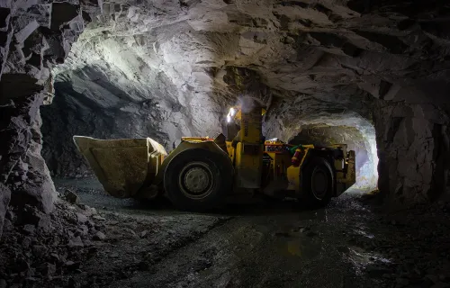 En lastmaskin arbetar i en upplyst gruvtunnel under jord. Den steniga tunnelmiljön och maskinen…