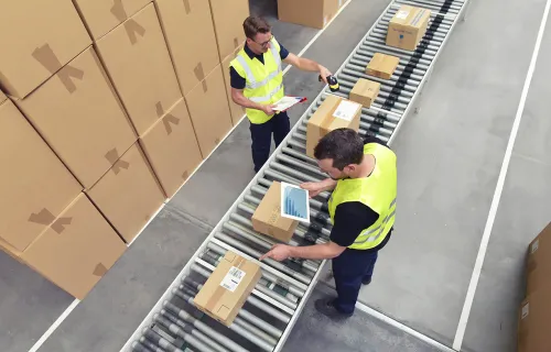 Två lagerarbetare i reflexvästar hanterar paket på ett rullband i ett lager. Kartonger är…