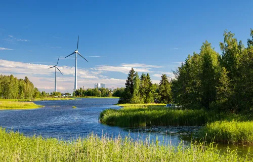 Vindkraftverk står vid en sjö omgiven av grön natur och träd under en klarblå himmel