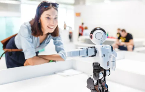 Kvinna står leende framåtlutad mot ett bord i ett öppet kontorslandskap och tittar på en robot