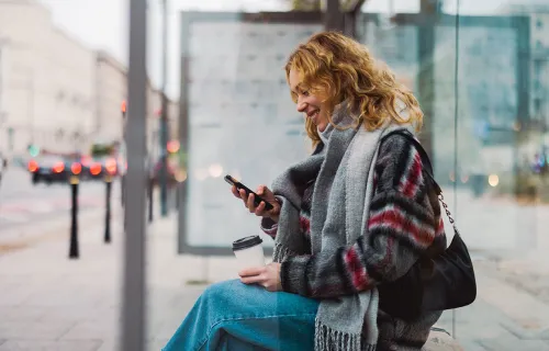 En kvinna sitter vid en glasvägg på en busshållplats i stadsmiljö och tittar leende på sin…