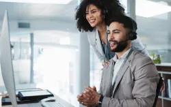 Two office workers wearing headsets look at a computer screen in a modern office