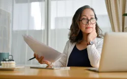 A woman holding paper looking at a laptop screen