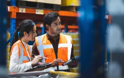 A warehouse worker and manager check stock and inventory using a digital tablet in a retail warehouse full of shelves containing goods.