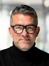 Porträtt av Anders Chrintz med grått hår, skäggstubb och svarta glasögon. Han bär en svart polotröja och tittar lugnt mot kameran, med en suddig kontorsmiljö i bakgrunden.