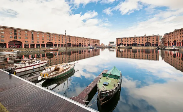  three boats at Albert Docks in Liverpool. The red buildings and cloudy sky are reflected in the water.
