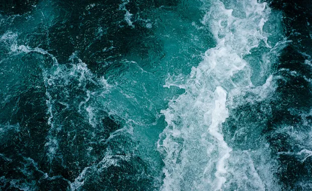 Närbild av havsvatten där vågor och skum bildar ett abstrakt mönster i turkosa och mörkgröna nyanser.