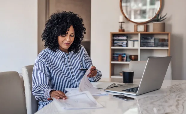 Kvinna sitter på sitt hemmakontor och tittar på papper framför sin laptop