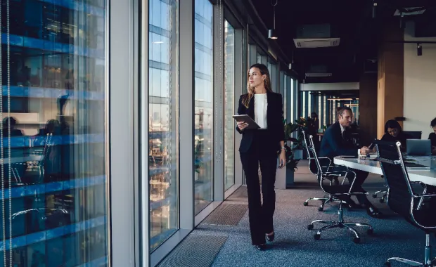 Responsable administrative en tenue formelle marchant dans un bureau avec une tablette numérique.