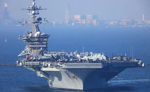 Aircraft carrier on calm blue waters with a cityscape in the background