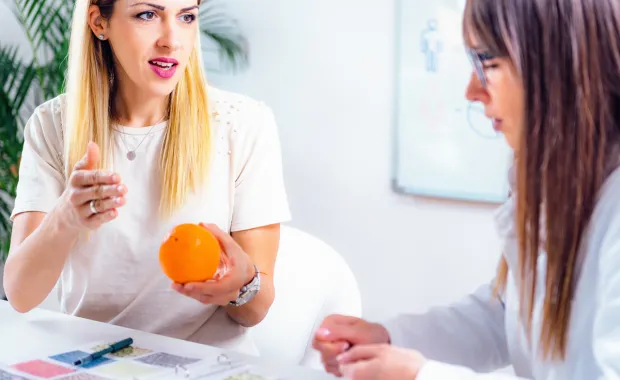 Två kvinnor sitter vid ett bord och pratar om levnadsvanor medan den ena kvinnan håller en apelsin i sin hand