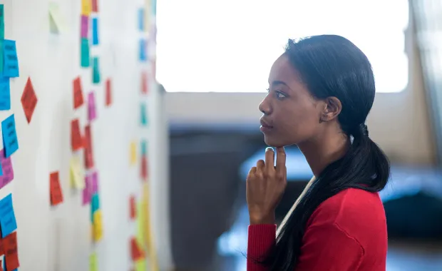 Kvinna står med handen under hakan och funderar medan hon tittar på post-it lappar i olika färger på en whiteboard