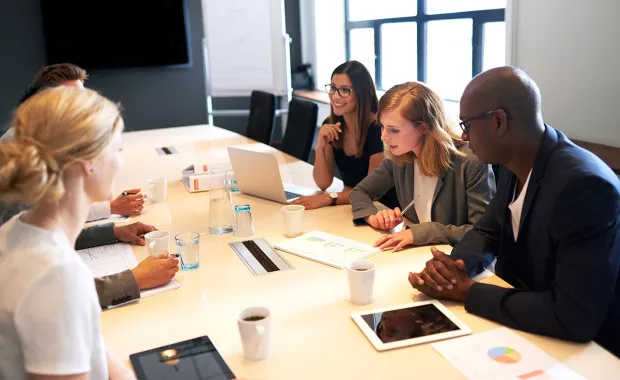 En grupp kollegor sitter runt ett konferensbord och diskuterar, med datorer, surfplattor och dokument framför sig.