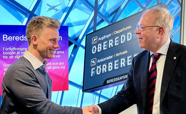 Två män skakar hand i en miljö med flygplatsskyltar i bakgrunden. Skyltarna visar avgångar och ankomster med namnen Örebro och Föreberg, och scenen signalerar samarbete eller ett officiellt möte.