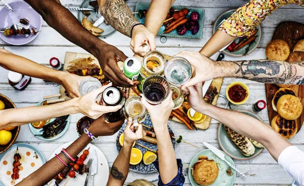 Viele verschiedene Menschen aus unterschiedlichen Nationen stoßen über einem mit Essen belegten Tisch an und symbolisieren so Diversität