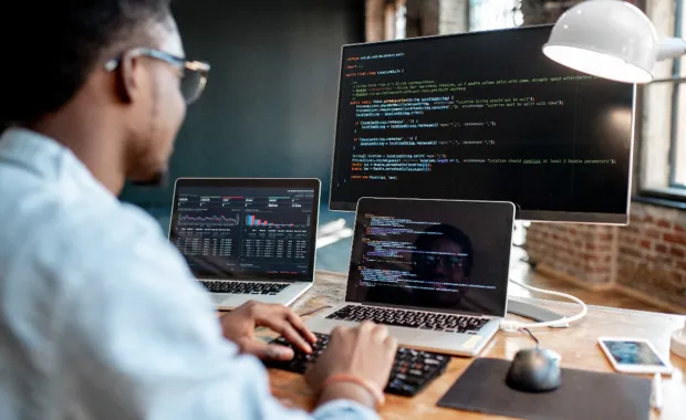 Person working at a desk with two laptops and a large monitor displaying code, data dashboards, and charts.