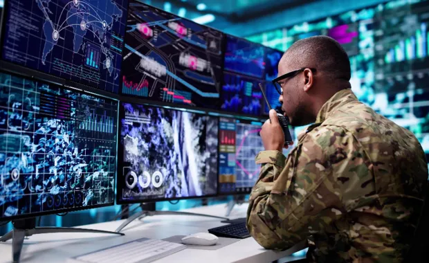 Dans une salle de contrôle, une personne en uniforme de camouflage militaire est assise à un bureau où s'affichent plusieurs écrans présentant des cartes, des images satellites et des graphiques de données.