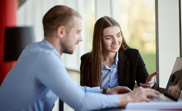 Två kollegor sitter tillsammans vid en bärbar dator och diskuterar, den ena visar något på skärmen.