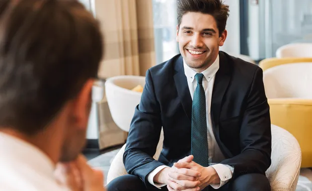 En man i kostym sitter leende och lyssnar på en annan kollega i en lounge