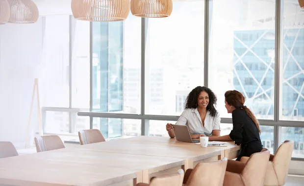 Två kvinnliga kollegor sitter och samtalar vid änden av ett konferensbord i ett stort rum med fönster ut mot storstadsvy