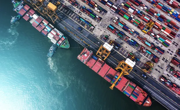 Flygfoto över en containerhamn med lastfartyg, kranar och färgglada containrar staplade längs kajen. Bilden visar logistik och internationell sjöfart från ovan.