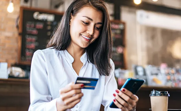 Frau sitzt in einem Cafe und hält in einer Hand ihre Kreditkarte und in der anderen Hand ihr Handy