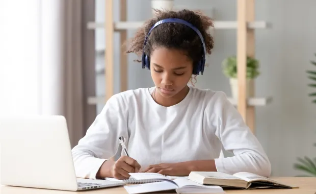 Ung student sitter vid sitt skrivbord hemma och lyssnar på undervisning samtidigt som hon tar noteringar i sitt block och i förgunden står laptopen uppfälld