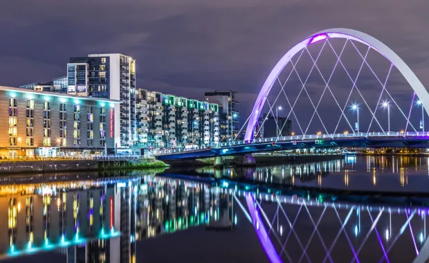 Illuminated white arch bridge spanning a river at night, with modern apartment buildings lining the waterfront
