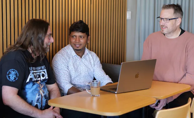 Tre kollegor sitter tillsammans vid ett bord och samtalar framför en laptop. De verkar engagerade i en diskussion i en avslappnad och modern arbetsmiljö, med en kaffekopp på bordet.