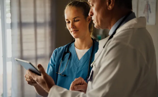 Två läkare, en i blå sjukvårdskläder och en i vit rock, granskar information på en surfplatta tillsammans.