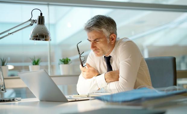 Man i vit skjorta sitter koncentrerat och läser på sin laptop på kontoret med sina glasögon i ena handen