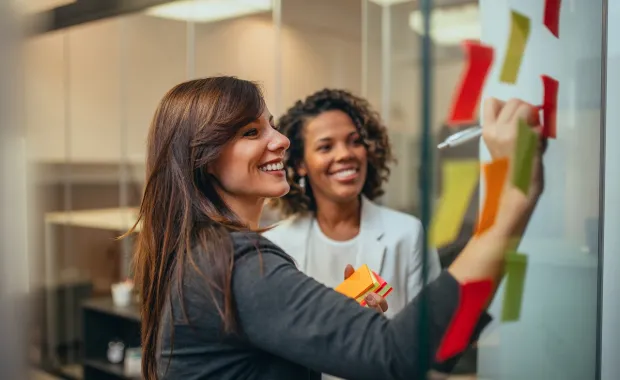 Två kvinnliga kollegor i ett agilt team står leende och brainstormar framför en whiteboard fylld av post-it lappar i olika färger